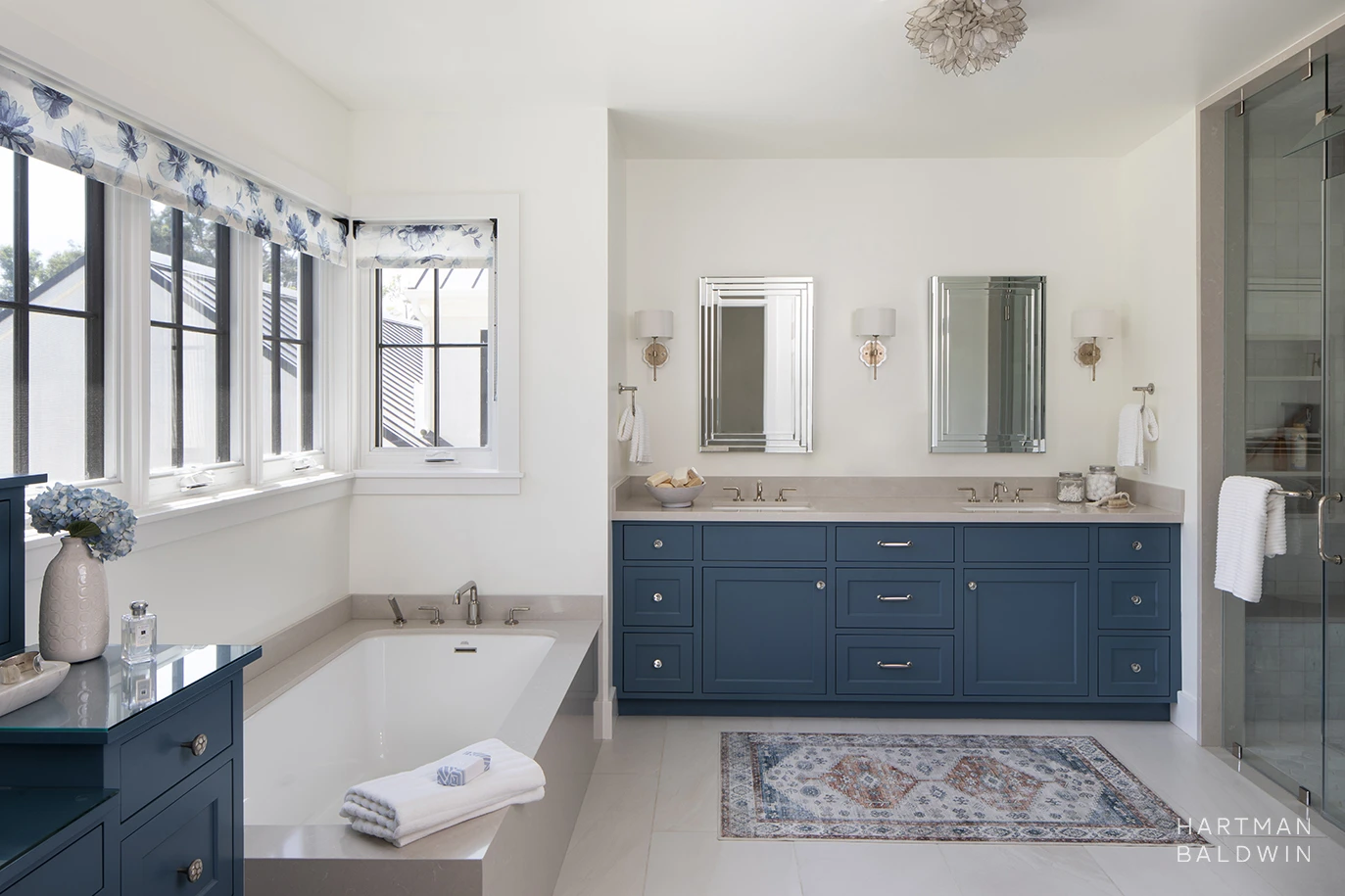 HartmanBaldwin Barrington Residence New Custom Modern Farmhouse Primary Bathroom - Hartman Baldwin New custom-designed master bathroom with walk-in glass shower, double sinks, large soaking tub, and built-in makeup vanity. The space features blue painted cabinetry, taupe stone surfaces, and floral-inspired lighting pieces