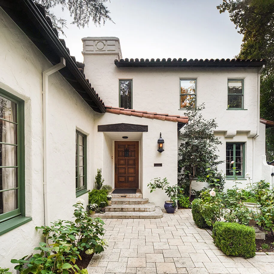 Front porch of a Spanish-style home with textured cream stucco walls, red-tile roof, green trim, and courtyard garden