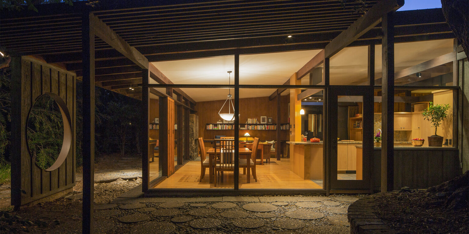 Remodeled Post-and-Beam Mid-Century Modern home after dark with warmly-lit kitchen, dining room, and living room visible through floor-to-ceiling windows