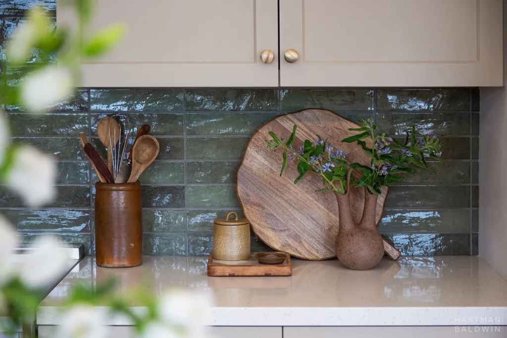 Kitchen detail with tan-painted cabinetry, brass hardware, light stone countertops, blue-green tile backsplash, and wood and ceramic kitchen accessories