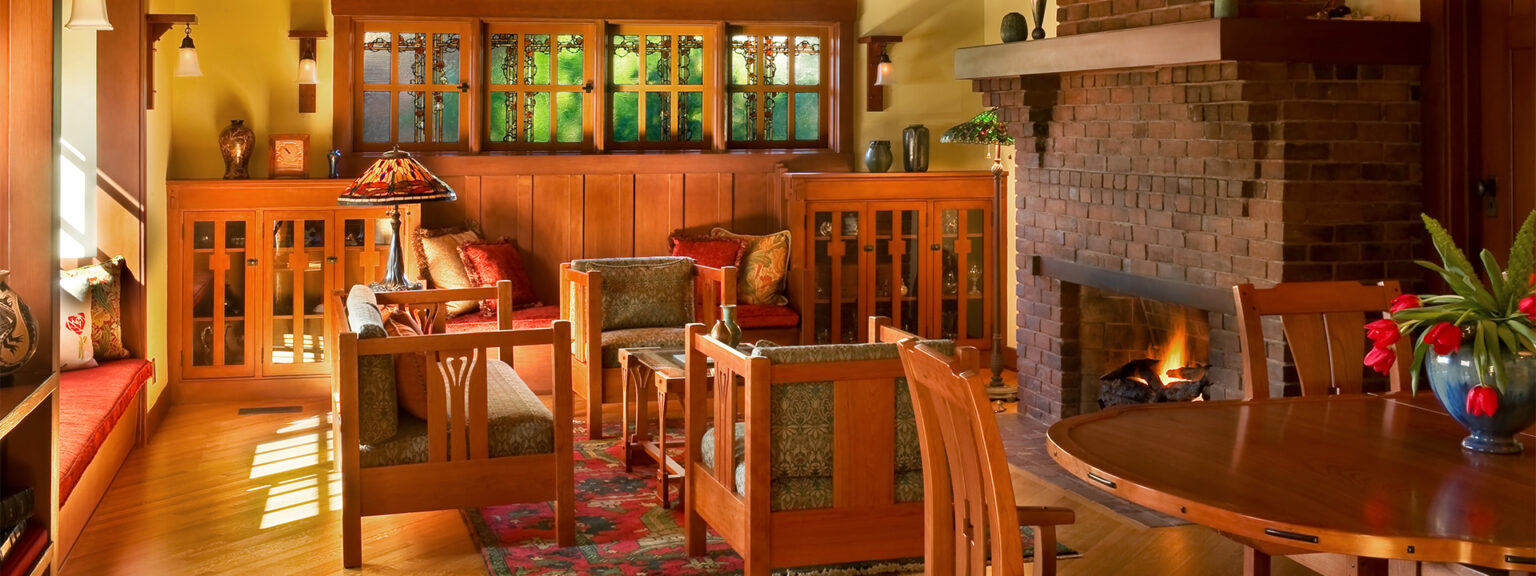 Craftsman-style living room at golden hour with brick fireplace, stained glass windows, built-in cabinetry, dining table, and window seat