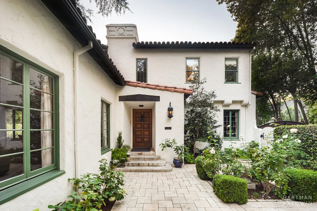 Front porch of a Spanish-style home with textured cream stucco walls, red-tile roof, green trim, and courtyard garden