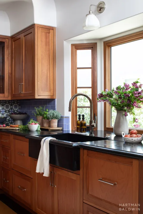 Remodeled Spanish-style kitchen with stained-wood custom cabinetry, blue backsplash tiles, and black soapstone countertops and sink in front of bay window