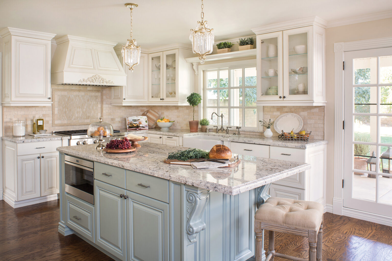 Remodeled traditional kitchen with intricate cream cabinetry, stone countertops, farmhouse sink, powder blue central island, and elegant glass pendant lights