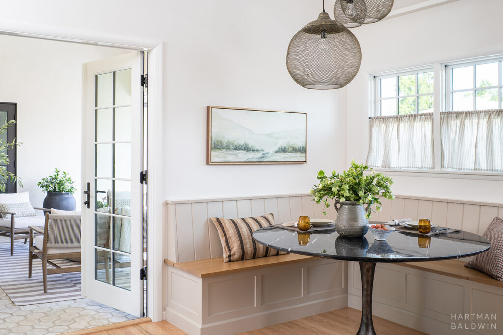 Corner breakfast nook with built-in banquette, round black stone table, and sculptural mesh pendant lights next to French doors open to adjacent California room patio