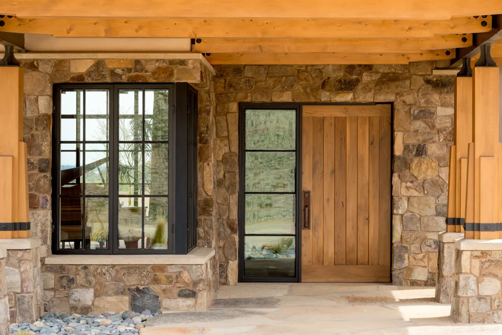 Front porch of new custom contemporary ranch-style California home featuring wood paneled front door, divided light steel windows, tumbled stone walls, and sturdy Alaskan yellow cedar pillars and columns