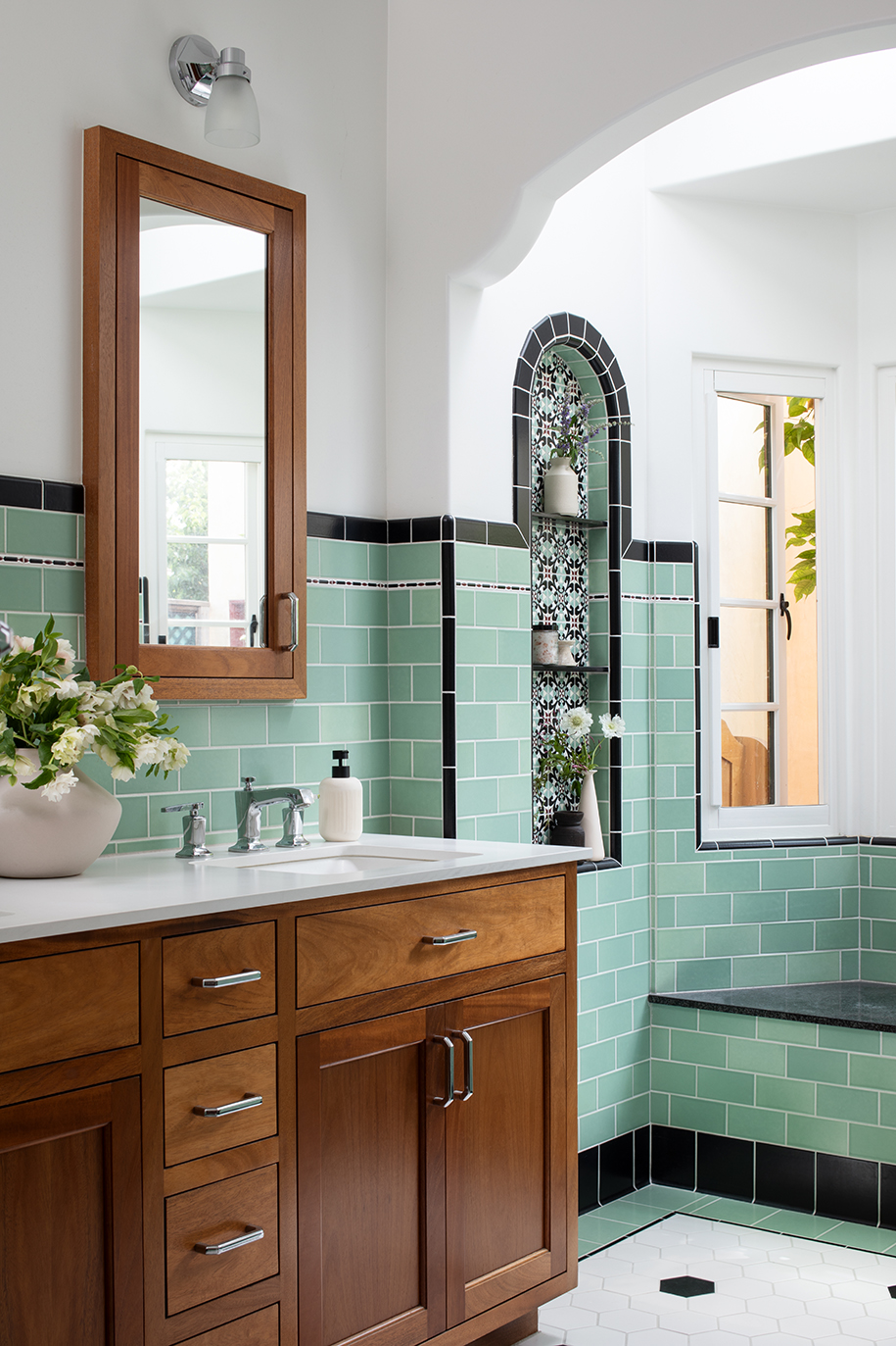 Spanish-style primary bathroom with warm mahogany cabinetry, seafoam-green wall tiles, decorative tiled niches, and a built-in window seat