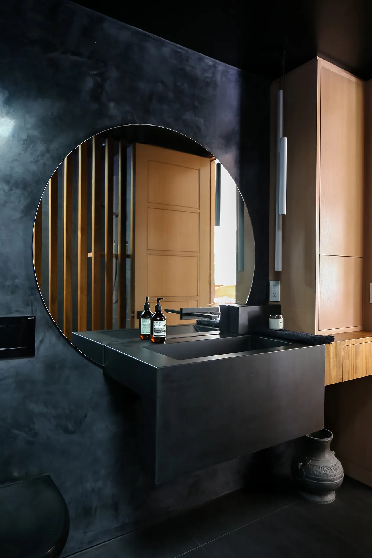 Remodeled bathroom interior design with black venetian plaster wall, large circular mirror, black concrete sink, and wood accents