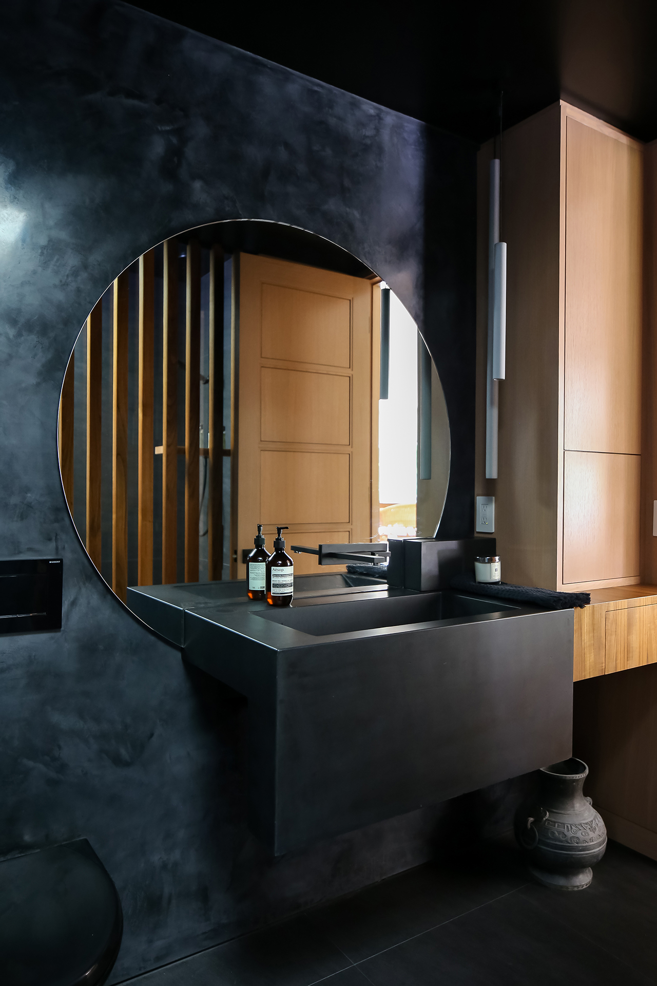 Remodeled bathroom interior design with black venetian plaster wall, large circular mirror, black concrete sink, and wood accents