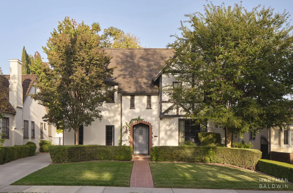 Two-story beige house with a brick arched doorway, framed by green hedges and trees on a sunny day.
