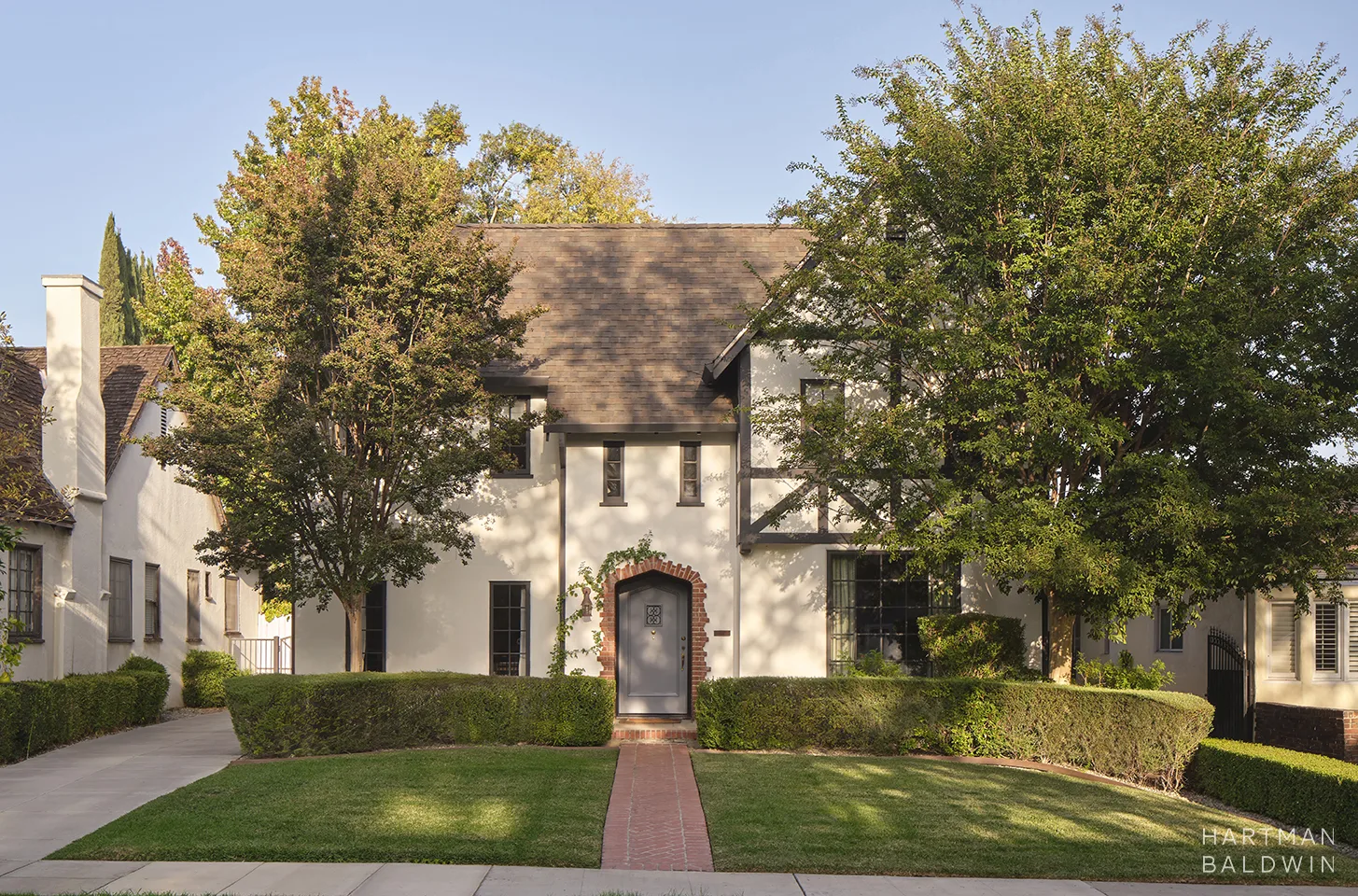 Two-story beige house with a brick arched doorway, framed by green hedges and trees on a sunny day.
