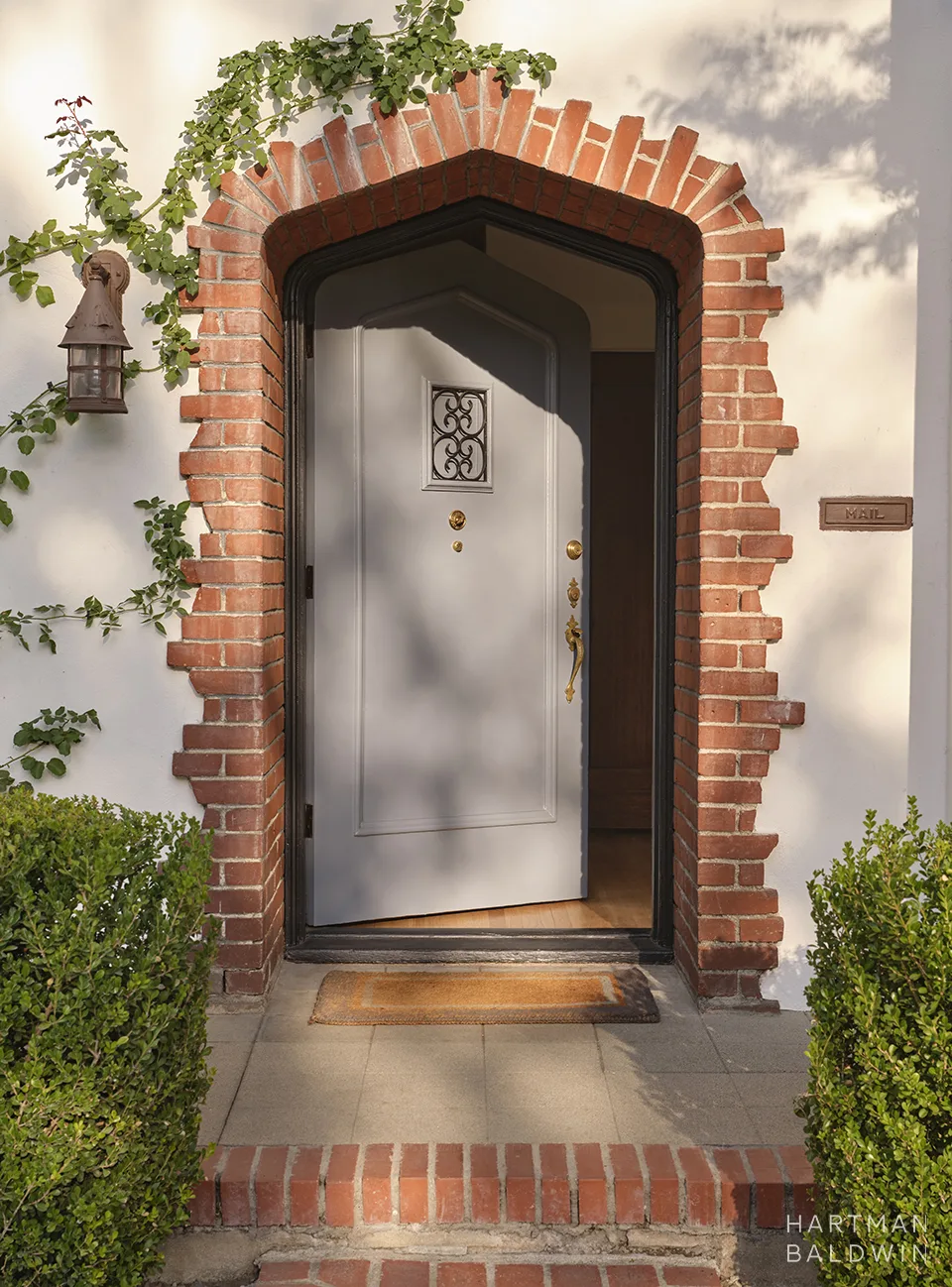 White front door with a decorative small window, set in a brick arched surround and flanked by a hanging lantern; greenery along theEntryway walls and a welcome mat outside.