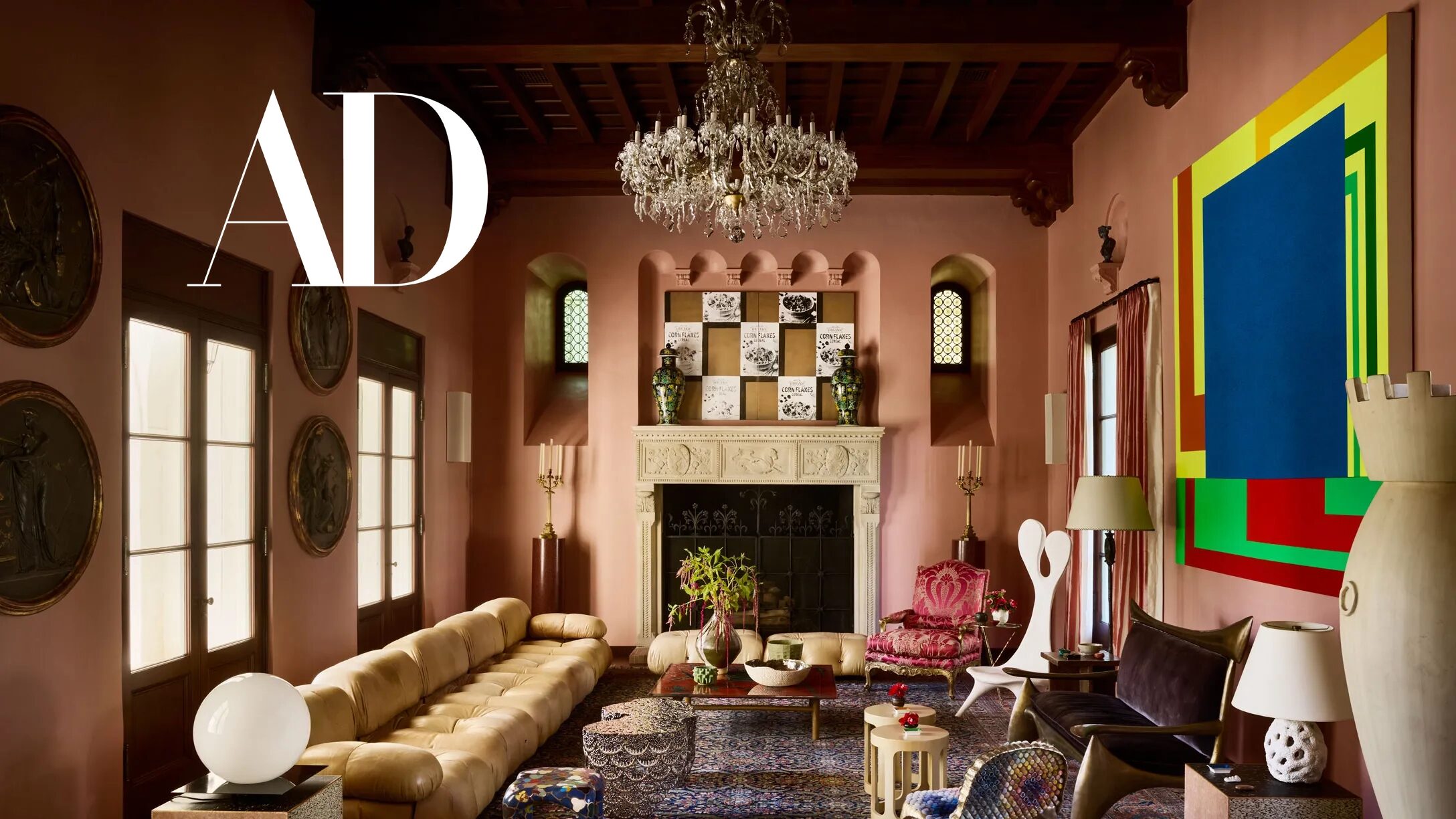 HartmanBaldwin Architectural Digest Feature Las Palmas Residence Hancock Park Spanish Style Estate Remodel Living Room - Hartman Baldwin Remodeled luxury historic double-height Spanish style great room with soft coral pink painted walls, dark wood ceiling beams, large carved stone fireplace, crystal chandelier, and a blend of traditional and modern furnishings and fine art selections. Architectural Digest Logo.