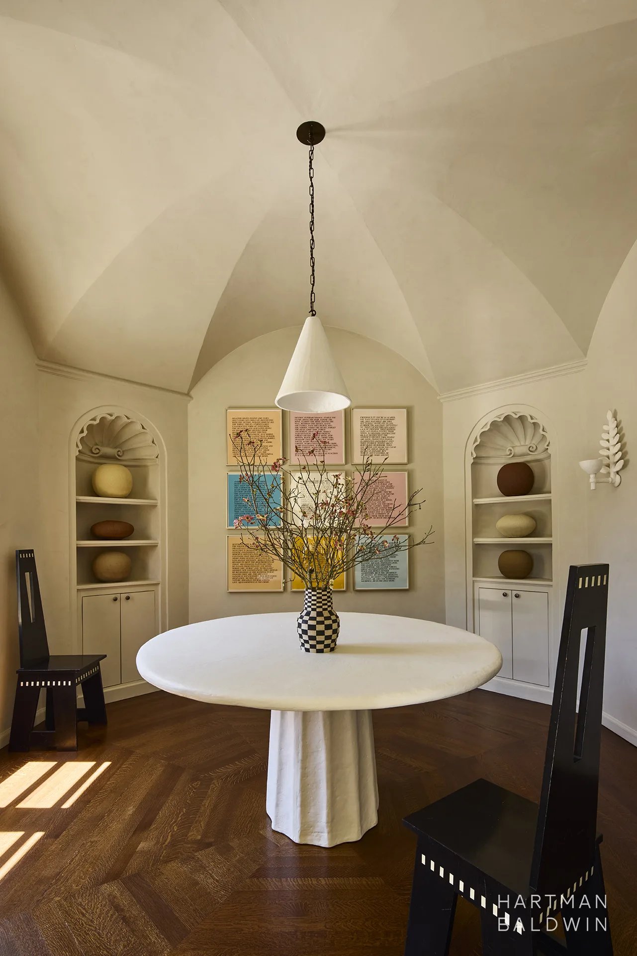 Remodeled Spanish-style breakfast room with a groin-vault ceiling, arched niches with carved details, framed art, and large round table with plaster texture that echoes the texture in the beige limewash walls and textured sculptural light pendant