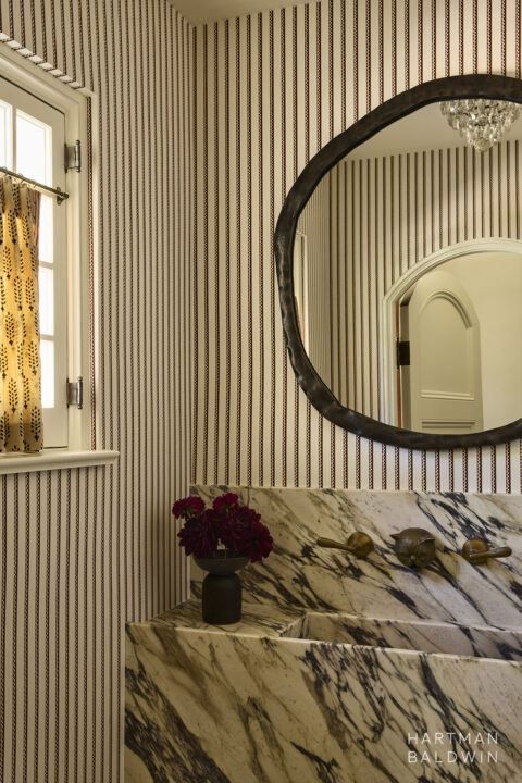 Bathroom interior design detail with striped wallpaper, custom bronze cat-shaped faucet, Calacatta Monet sink console, and asymmetric mirror.