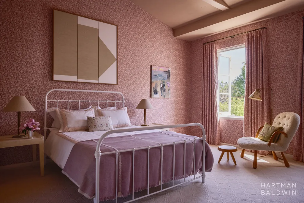 Feminine bedroom with vibrant pink and white floral patterned wallpaper and matching drapes, soft pink-painted vaulted ceiling, bed with pink bedding, comfortable white chair with lamp, and abstract art