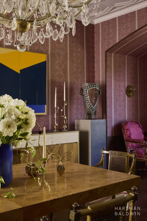 Historic home dining room with patterned red and white wallpaper, modern and historic furnishings and fine art, and crystal chandelier. A burlwood dining table is paired with vintage chairs from Queen Victoria's collection.