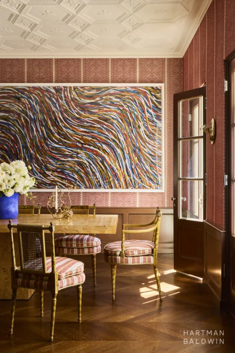 Historic home dining room redesign with patterned red and white wallpaper, ornate ceiling detailing, a large abstract painting, and hardwood flooring. A door to the outside lets in natural light, and a burlwood dining table is paired with vintage chairs from Queen Victoria's collection.