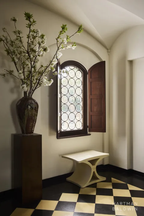 Spanish-style entry hallway detail. Arched window with dark-stained wood framing and circular glass detailing, white sculptural stool, and large floral arrangement in vase on a black pedestal. Cream and black checkered tile flooring.