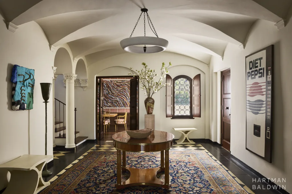 Remodeled historic Spanish-style home entryway with groin-vault ceiling details, sculptural light pendant, and fine art selections. The colorful dining room beyond is seen through a doorway.
