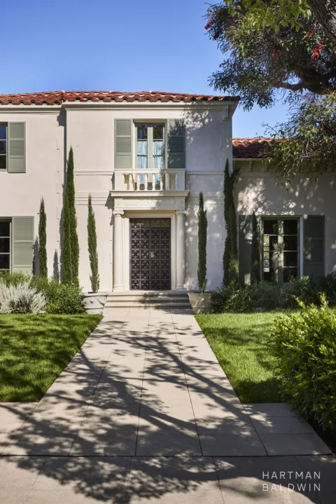 Hancock Park Los Angeles Spanish Colonial Revival-style estate front exterior with cream stucco walls, sage green window shutters, red-tile roof, traditional columns, cypress trees, and patterned wrought-iron front gate.