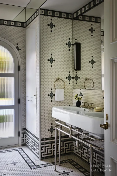 Remodeled Spanish-style pool bathroom with patterned cream and white tile on the floor and walls, a white and silver double sink, and arched door with fluted glass