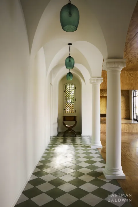 Remodeled Spanish-style hallway with white plaster columns and arches, groin-vault ceilings, green and white tile flooring, green glass light pendants, an arched stained glass window, and a vintage chair. To the right of the hallways is a luxury wood-paneled home gym with arched glass windows that overlook the backyard.