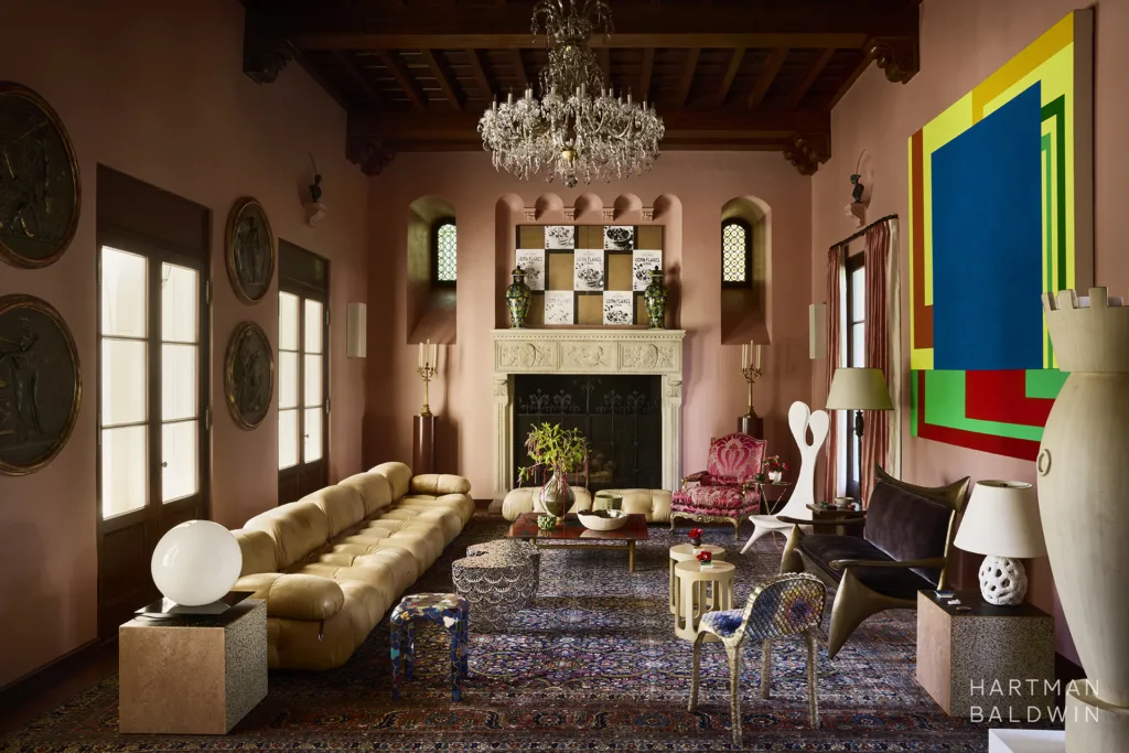 Remodeled luxury historic double-height Spanish style great room with soft coral pink painted walls, dark wood ceiling beams, large carved stone fireplace, crystal chandelier, and a blend of traditional and modern furnishings and fine art selections.
