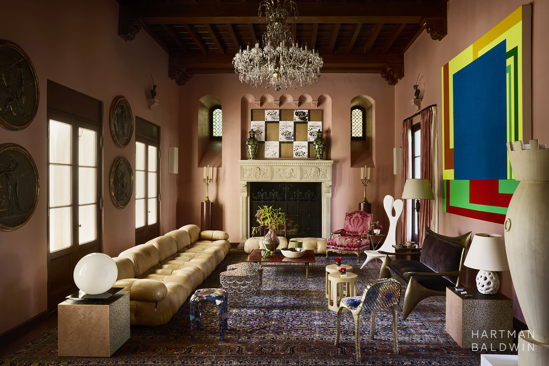 Remodeled luxury historic double-height Spanish style great room with soft coral pink painted walls, dark wood ceiling beams, large carved stone fireplace, crystal chandelier, and a blend of traditional and modern furnishings and fine art selections.