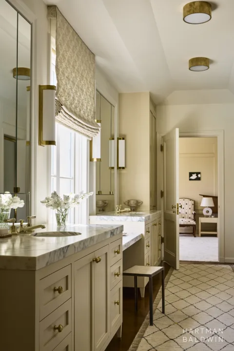 Remodeled luxury primary bathroom. The built-in cabinetry includes two sink vanities, a space to sit in front of a large window, and a floor-to-ceiling linen closet. The cabinetry features white stone countertops with grey veining and gold-tone hardware.