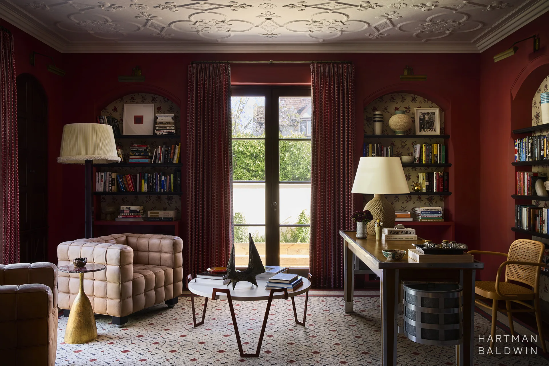 Moody vintage-inspired office with deep red walls and curtains, ornate ceiling detailing, arched niche bookshelves, comfortable seating, and wooden desk.