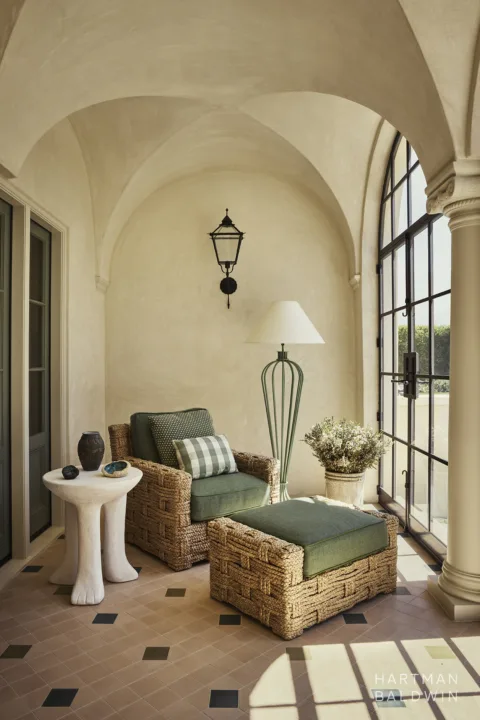 Spanish-style sunroom with columns framing tall arched windows, groin-vault ceiling details, limewash walls and ceiling, tiled floor, and comfortable lounge chair with abstract side table and lamp.